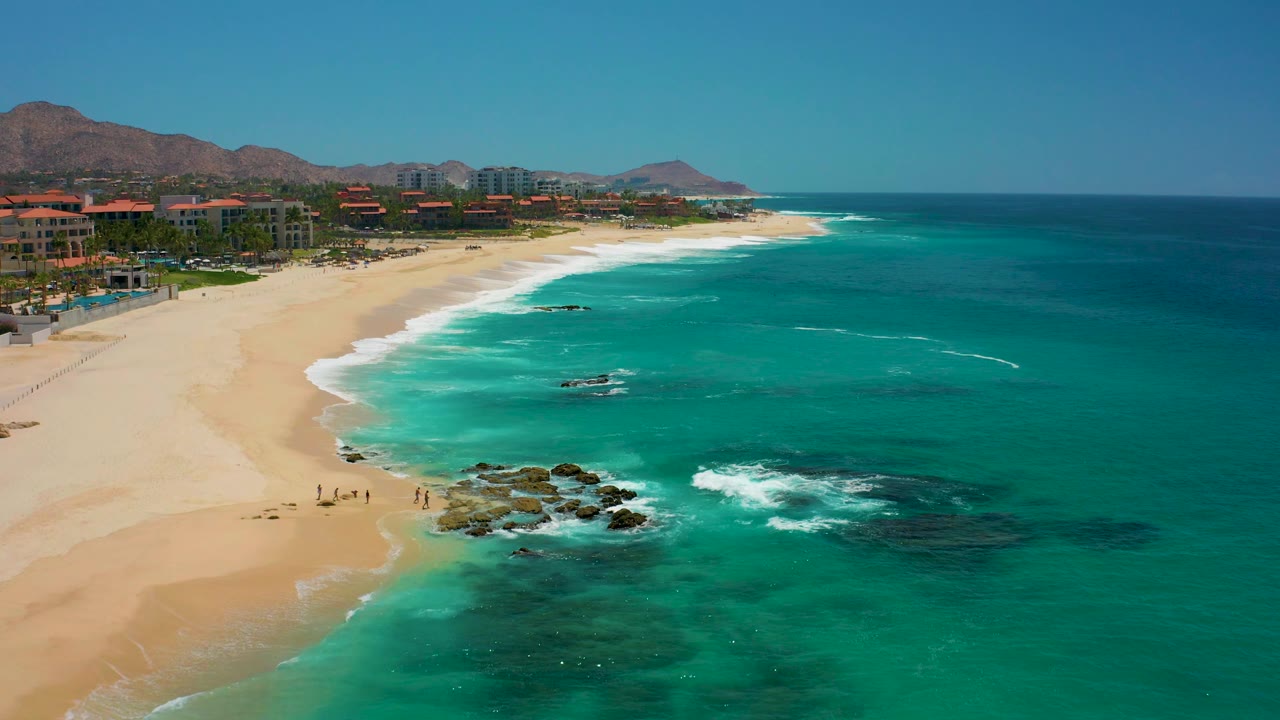 Aerial view at Dreams Los Cabos in Baja California Sur showing a broad sandy beach, turquoise ocean, rocks in the surf, small people on the sand, beachfront buildings, and mountains in the distance.