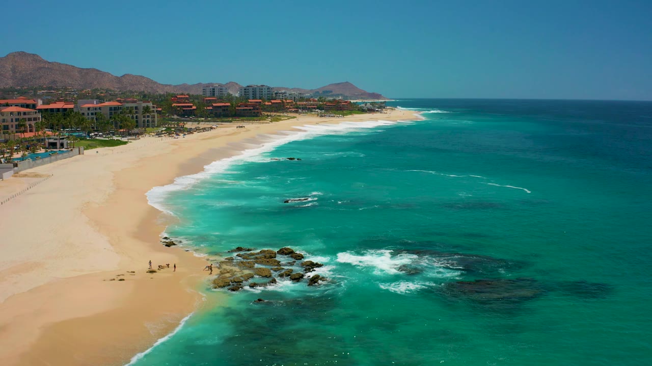 Aerial frame at Dreams Los Cabos showing the beach stretching into the distance, a bright turquoise bay, white water around the foreground rocks, small beachgoers on the sand, and low mountains behind the buildings.