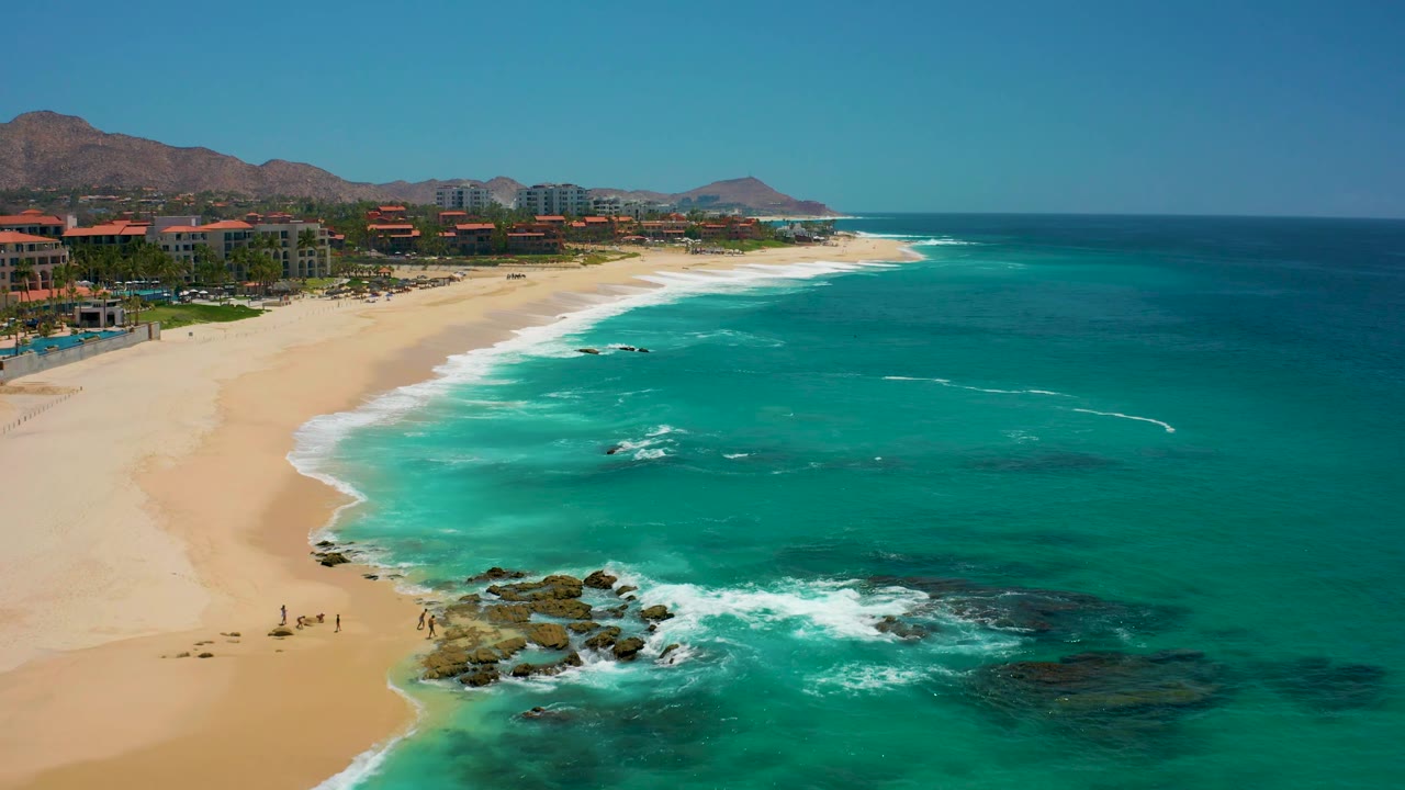 Beach and ocean seen from above with rocks, foam, people, buildings, and mountains.