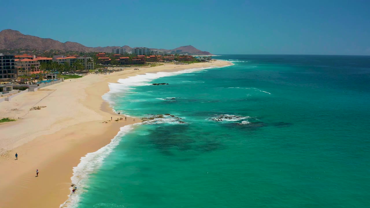 Wide aerial shoreline with waves, beachgoers, rocks, resort buildings, and hills.