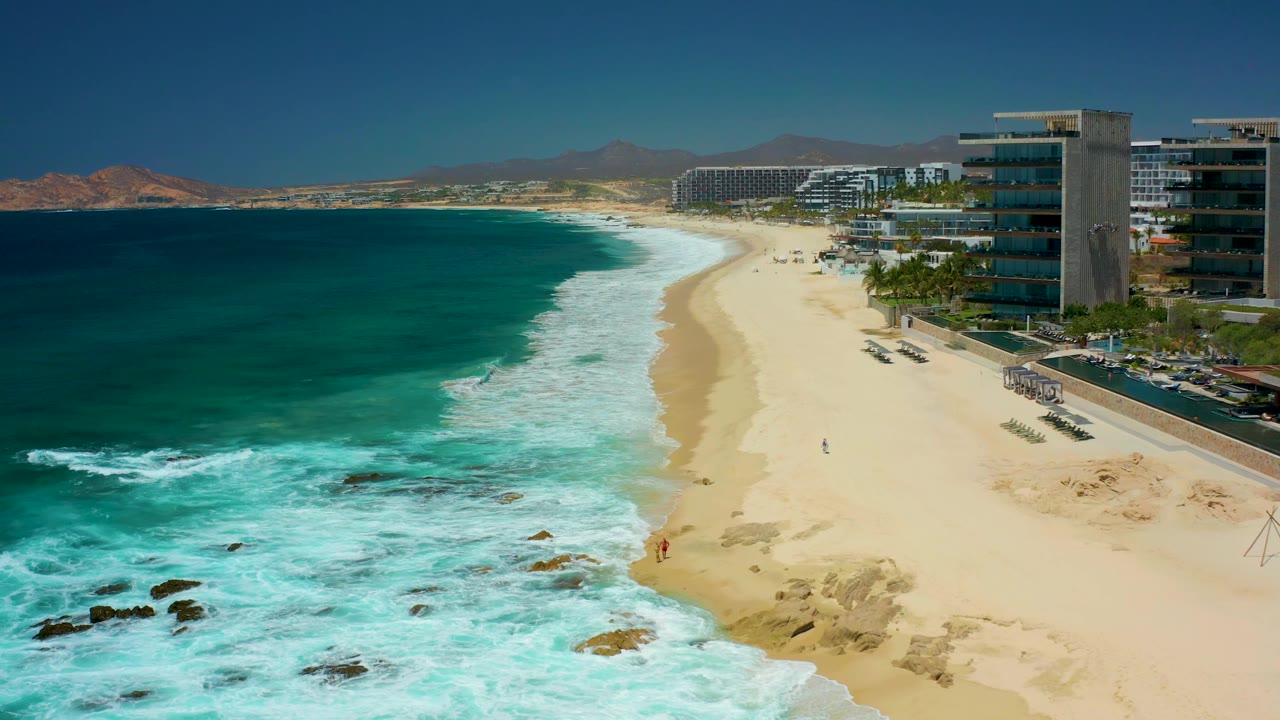 Waves roll toward a sandy beach beside coastal buildings and a beachfront road.