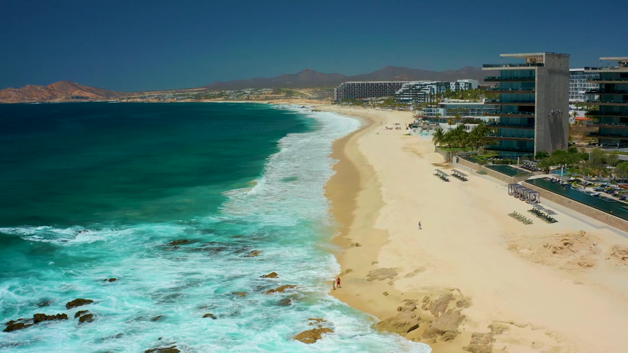 The shoreline at Dreams Los Cabos shows foamy turquoise water spreading over the sand, beachfront buildings to the right, a road with vehicles, and small figures near the waterline.