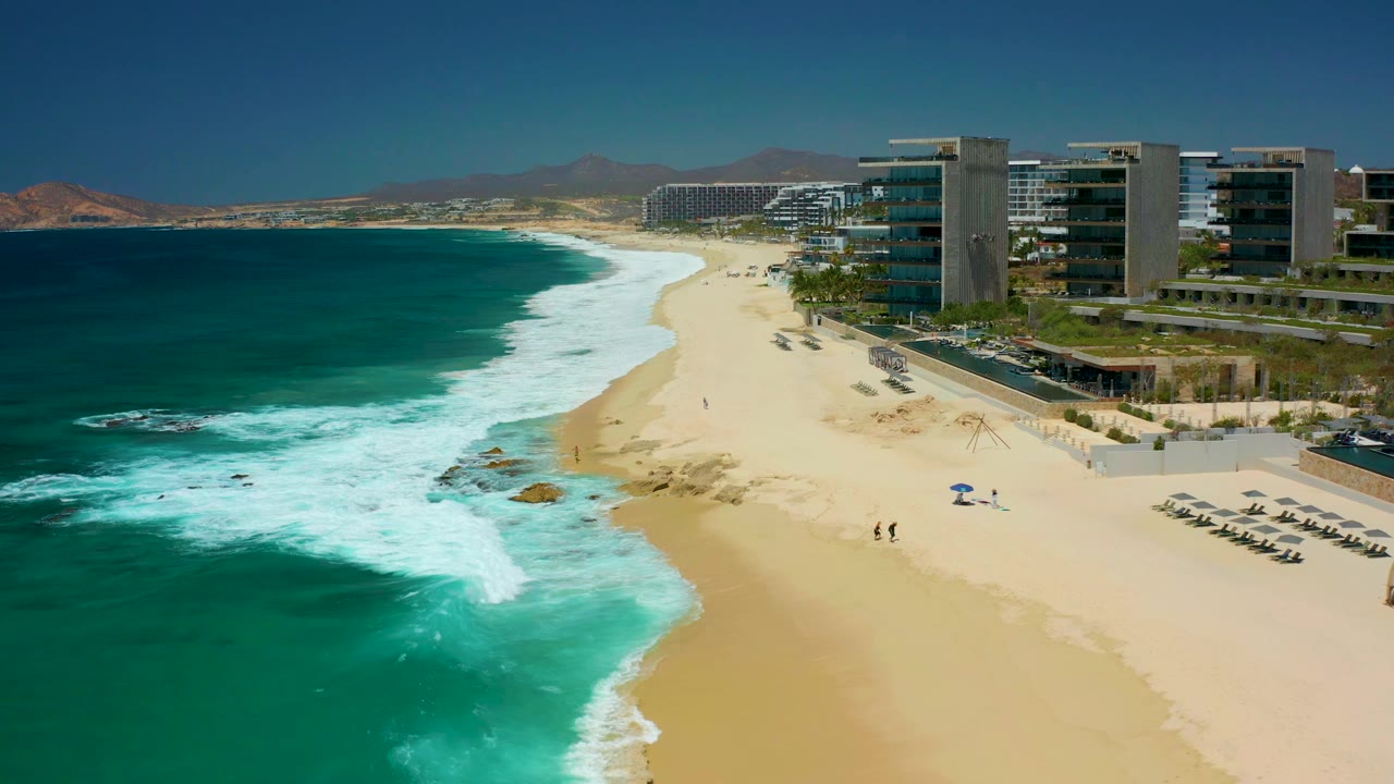 Aerial frame at Dreams Los Cabos showing turquoise water breaking onto a wide sandy beach, resort buildings on the right, distant hills, beach chairs, a blue umbrella, and small visible people near the shore.