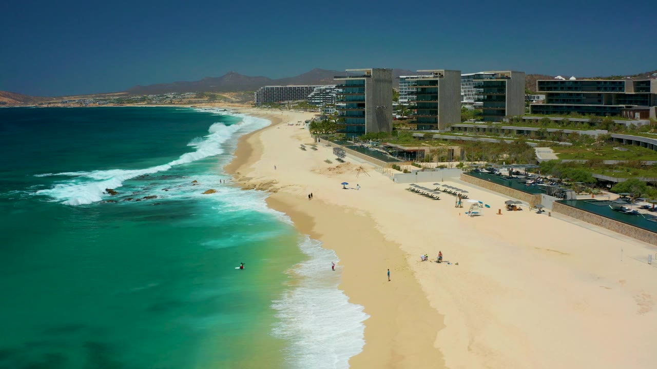 Aerial view at Dreams Los Cabos, Baja California Sur, with turquoise surf rolling onto a wide sandy beach, scattered people, and modern coastal buildings along the right side.
