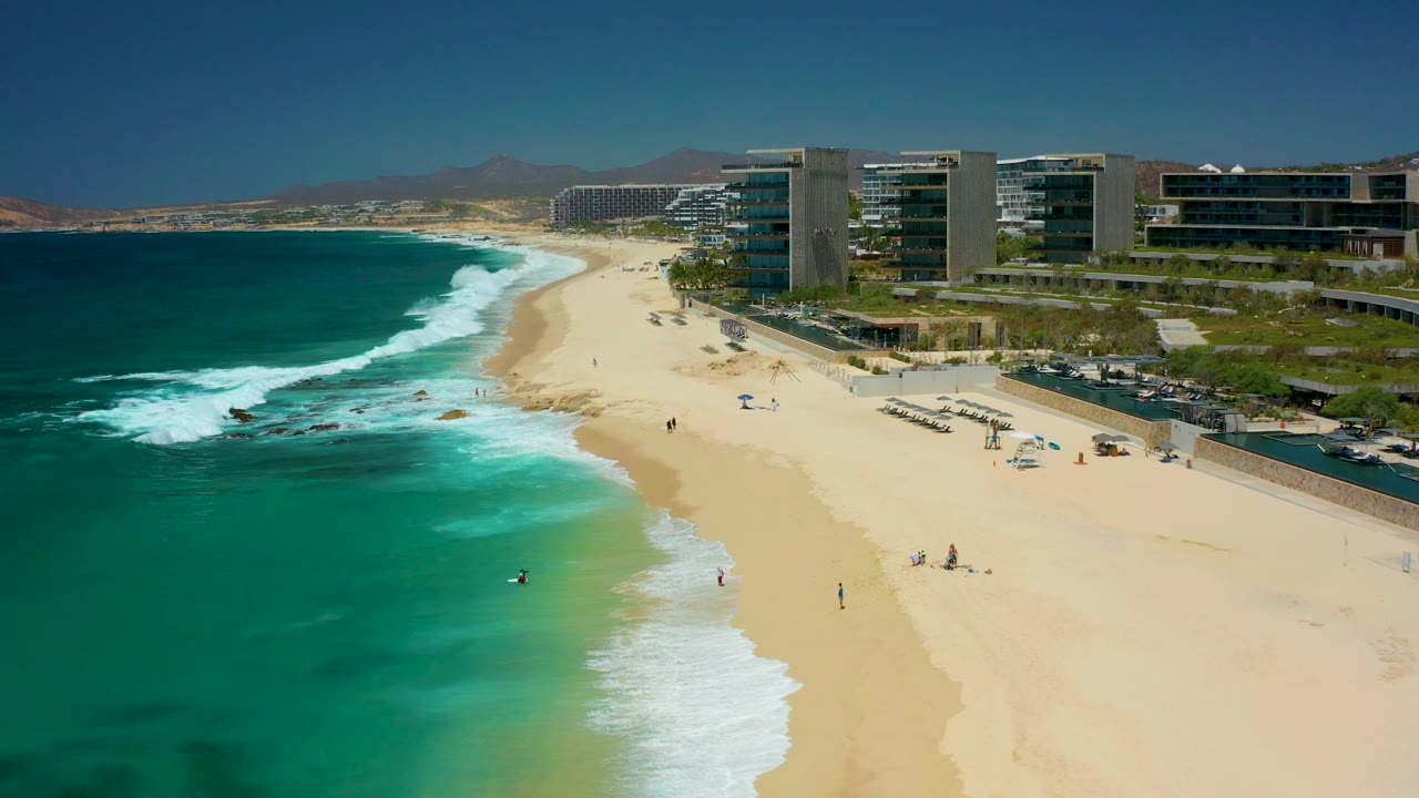 Turquoise surf breaks along a sandy beach with beachfront buildings nearby.