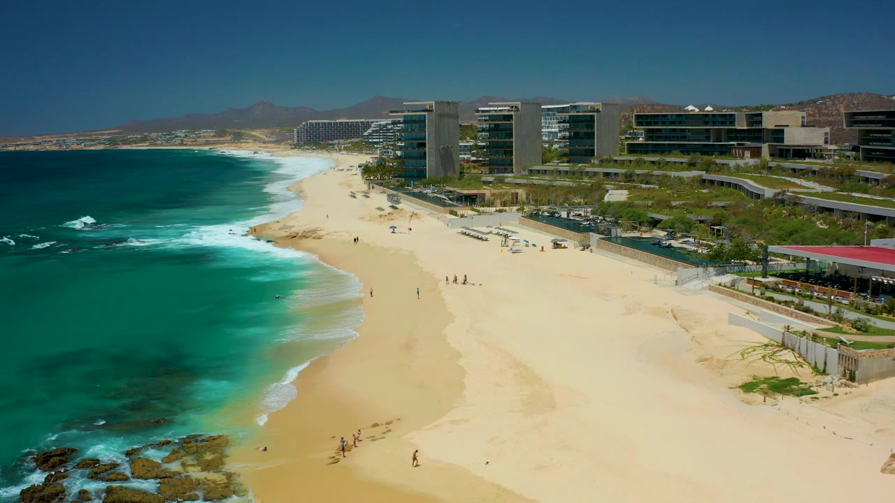 Aerial shoreline view with waves curling toward sand and resort buildings behind the beach.