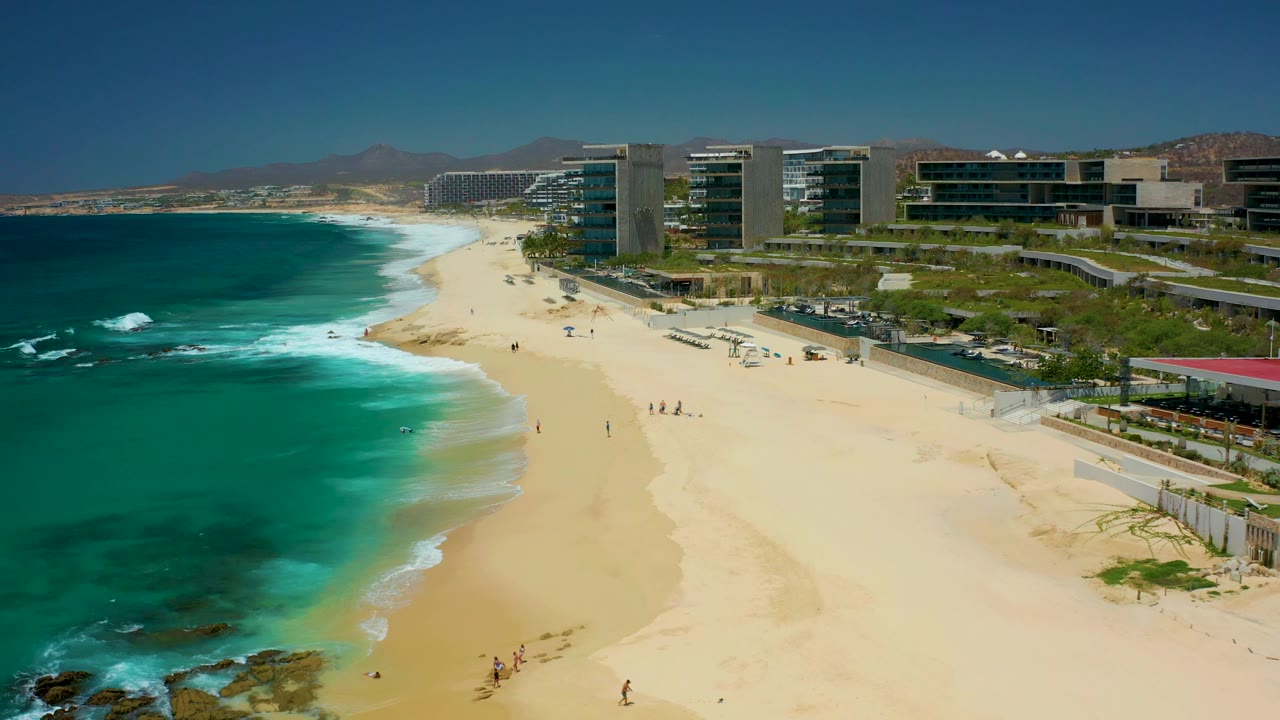 At 0:02, the elevated Dreams Los Cabos view shows clearer open sand in the lower right, turquoise water along the left edge, scattered beachgoers, and resort buildings across the upper right.