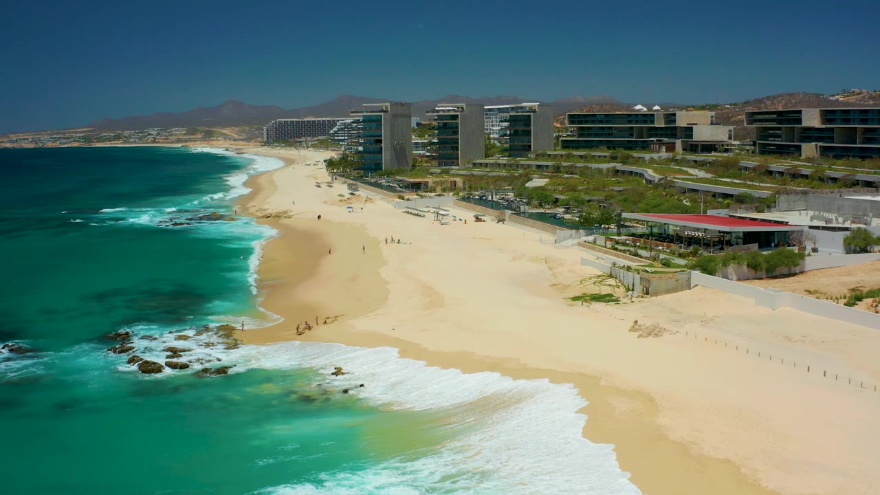 Waves roll toward a wide sandy beach beside modern coastal buildings.