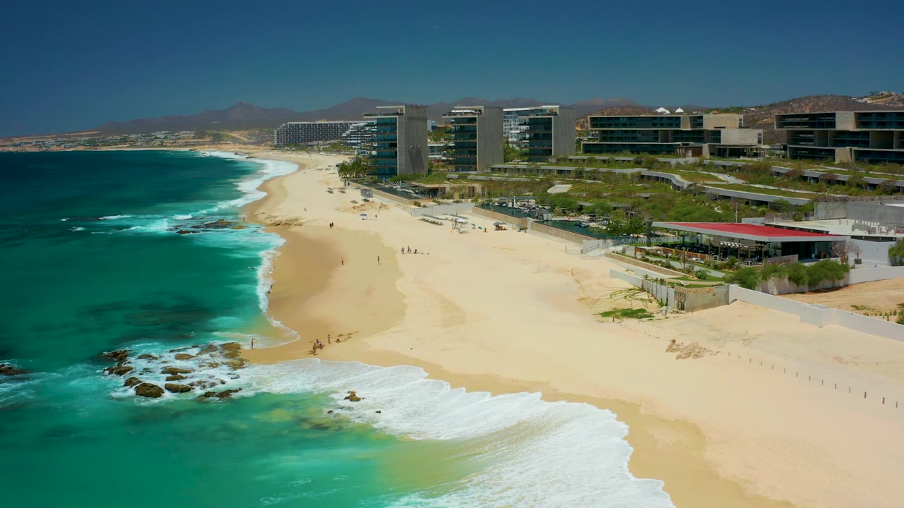 Aerial still over Dreams Los Cabos showing a broader band of foam near the lower center, small figures spread along the sand, beachfront structures to the right, and mountains beyond the resort area.