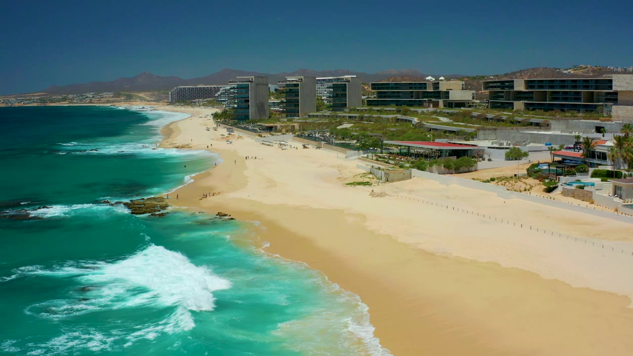 Aerial view of turquoise waves breaking beside a sandy beach and coastal buildings.
