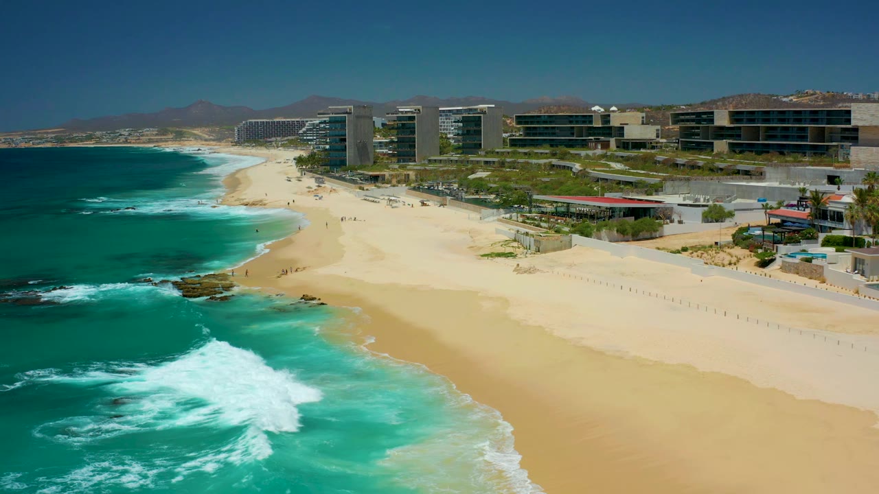 At 0:01, the Dreams Los Cabos shoreline remains wide in frame as a white wave patch spreads near the lower left, with beach sand, coastal buildings, distant hills, and no visible people.