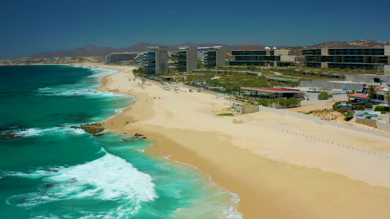 At 0:02, white foam gathers farther along the left shoreline while pale sand opens across the center, with Dreams Los Cabos coastal buildings in the background and no visible people.