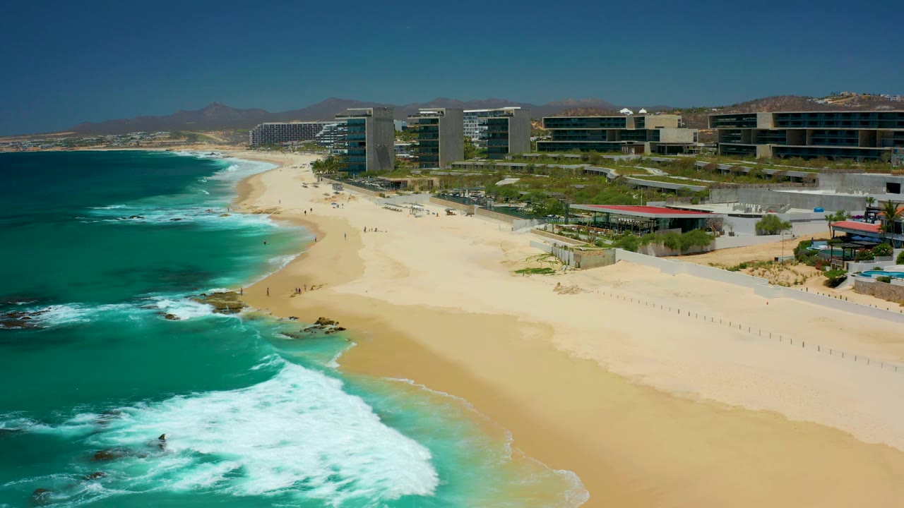 At 0:03, stronger white surf fills the lower-left foreground, the sandy beach curves into the distance, coastal buildings sit above the shore at Dreams Los Cabos, and no visible people appear.