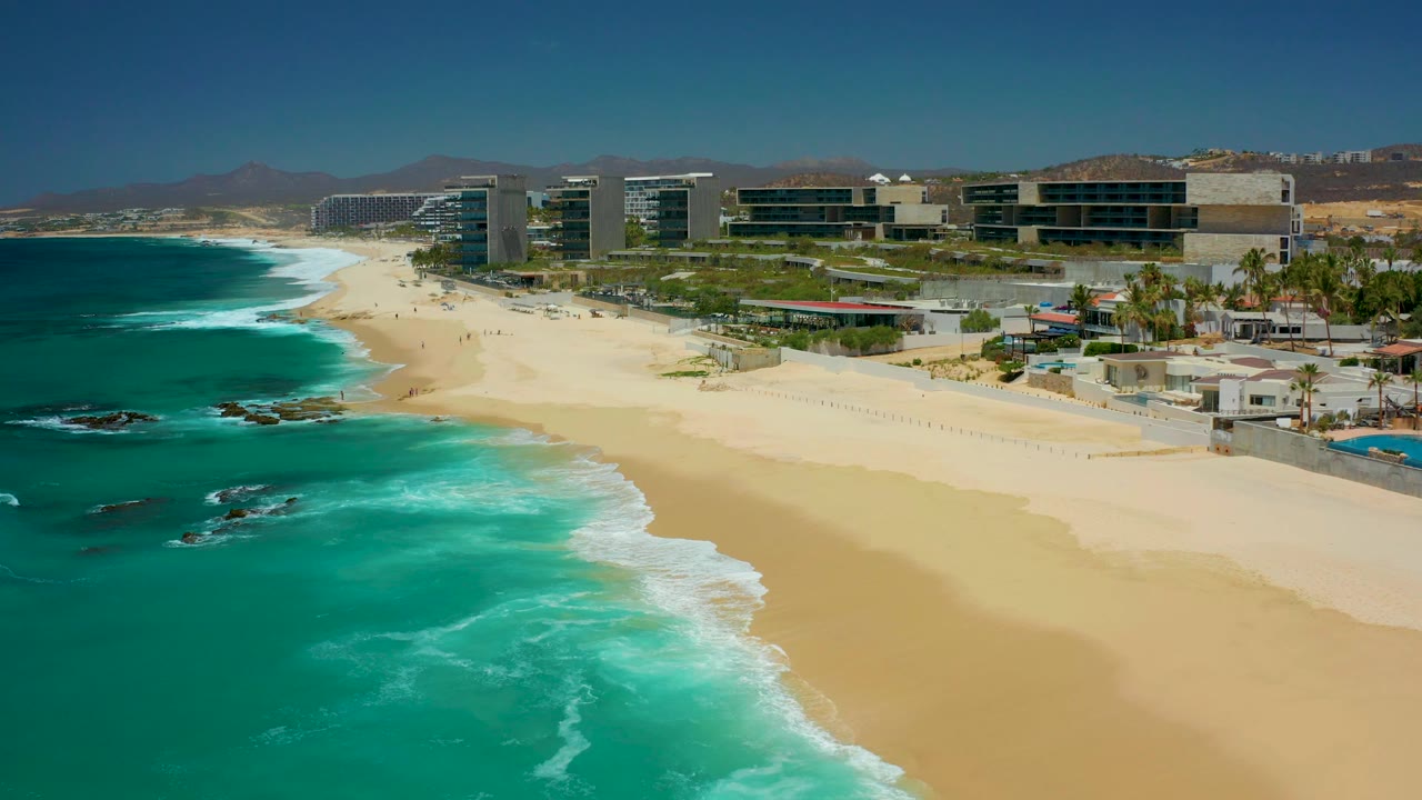 At Dreams Los Cabos in Baja California Sur, turquoise water curves toward a pale sandy beach with modern coastal buildings and distant small beachgoers visible.