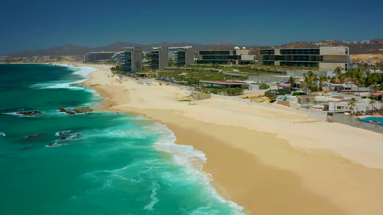 Turquoise surf approaches a wide beach below modern coastal buildings.
