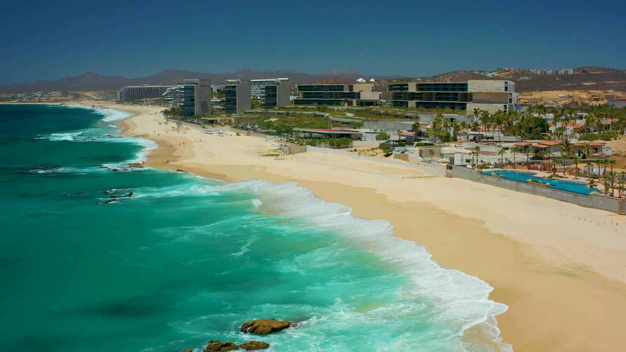 Opening aerial frame at Dreams Los Cabos shows turquoise water and white surf meeting a broad sandy beach, with resort buildings, palms, and distant people along the shore.