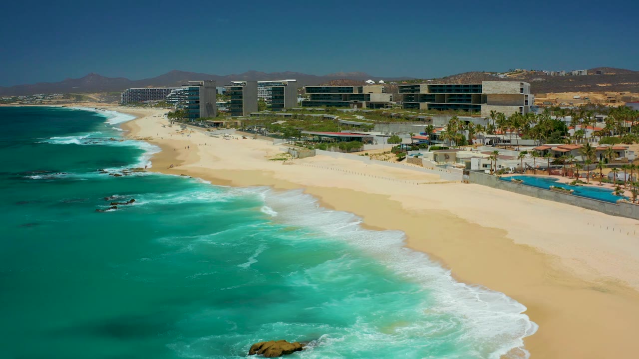 At 0:02, the aerial view holds the turquoise ocean on the left, a long curve of pale sand on the right, and Dreams Los Cabos buildings backed by dry hills.