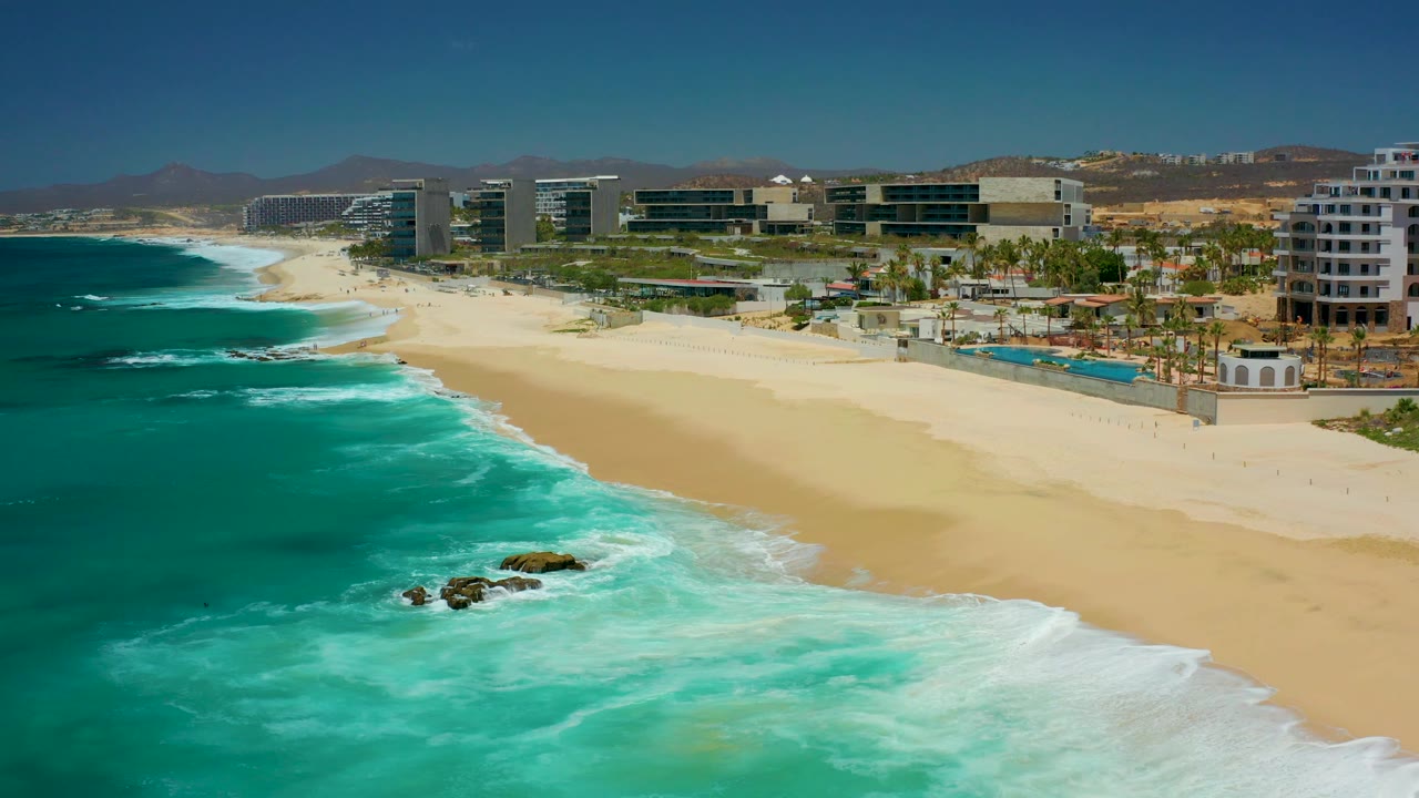Aerial view at 0:01 over Dreams Los Cabos, where foamy turquoise waves reach the sand while resort buildings, pools, palm trees, hills, and a clear sky fill the background with no visible people.