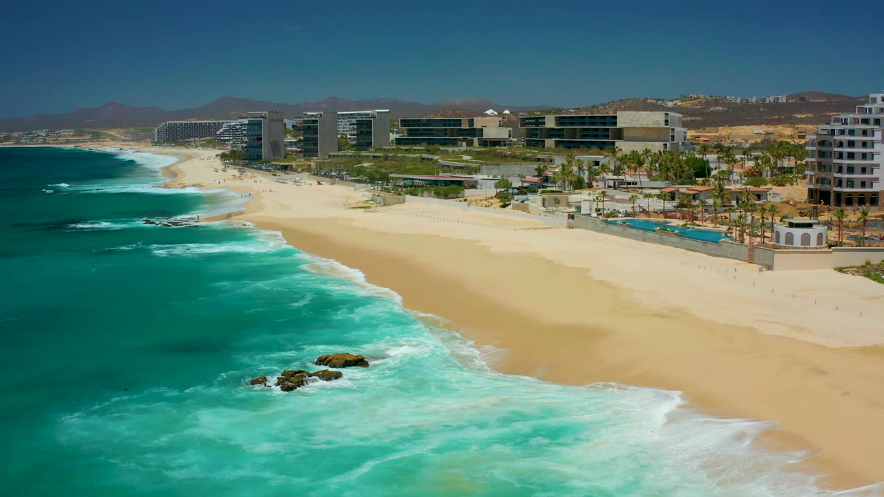 Aerial frame at Dreams Los Cabos showing a slightly shifted shoreline view, with white surf curling around rocks, pale sand along the right side, resort pools and palms behind it, and no visible people.