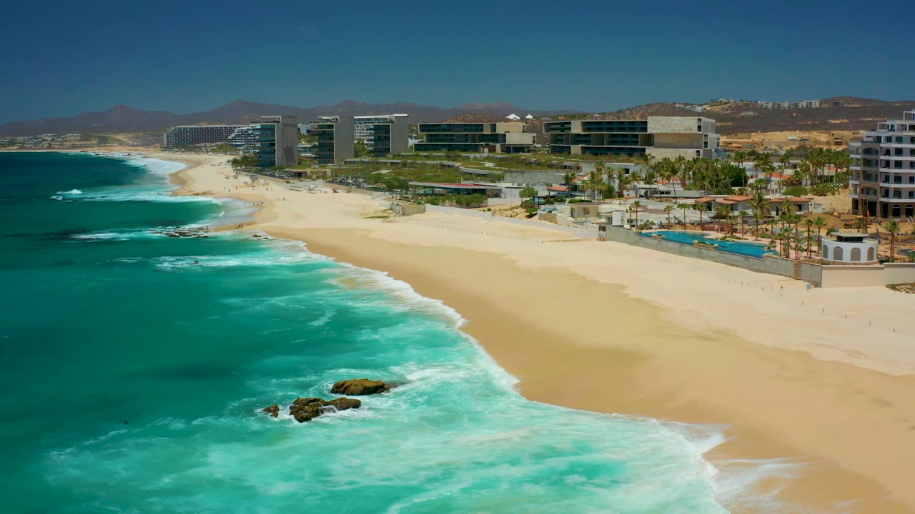 Aerial frame at 0:03 over Dreams Los Cabos, with darker turquoise water at left, foamy waves crossing the sand, rocks near the lower frame, palm-lined pools behind the beach, and no visible people.