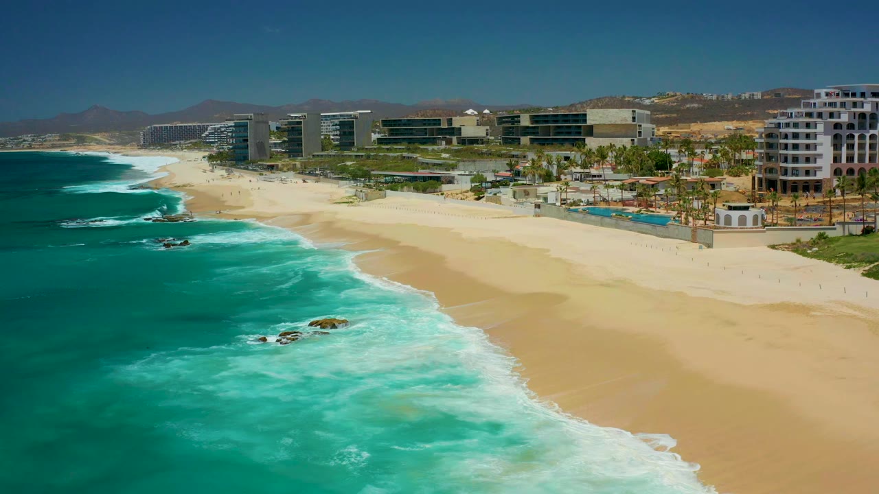 Opening aerial frame of Dreams Los Cabos with turquoise surf on the left, pale sand on the right, resort buildings and palms in the background, and no visible people.