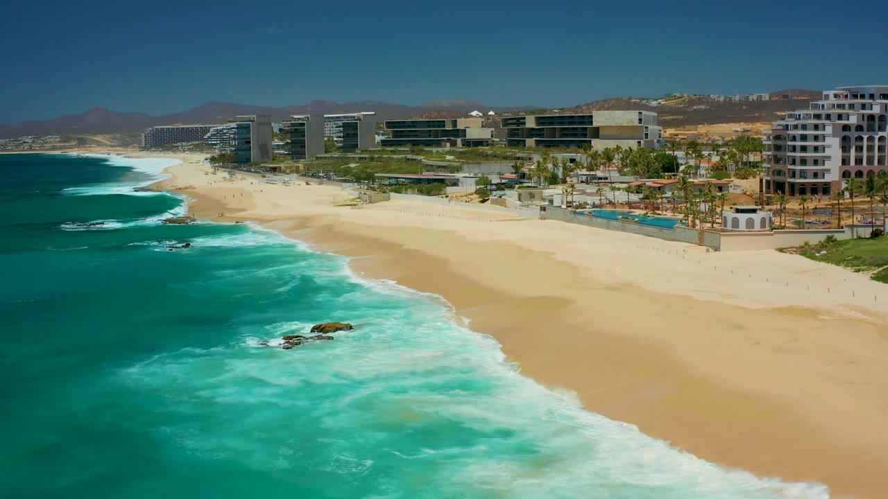 Aerial frame at 0:02 with the ocean occupying the left half, a broad sandy beach curving toward resort buildings, low hills beyond, and no visible people.