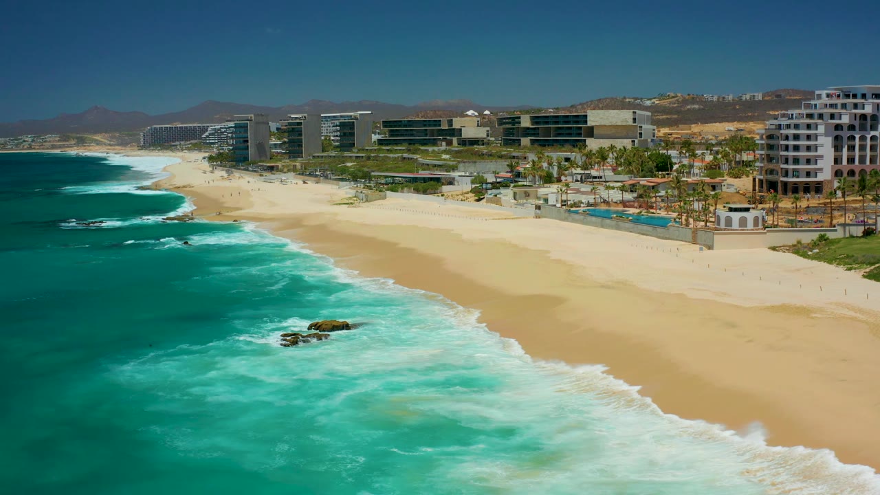 Aerial shoreline view with surf, rocks in the water, sand, and resort frontage.