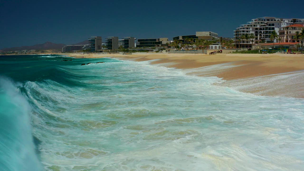 A turquoise wave approaches tan sand at Dreams Los Cabos, with beachfront buildings in the background and no visible people.