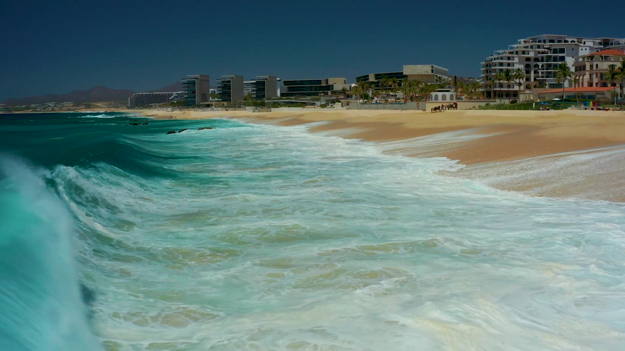 A larger turquoise swell rolls along the left side of the frame toward the sandy Dreams Los Cabos shoreline, with coastal buildings beyond and no visible people.
