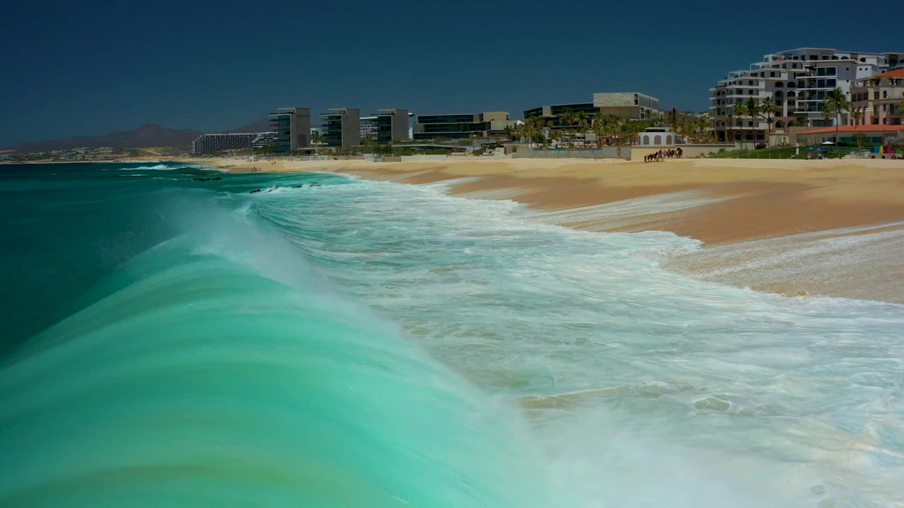 A foamy turquoise crest fills the foreground as it moves toward the Dreams Los Cabos sand, with resort buildings across the upper background and no visible people.