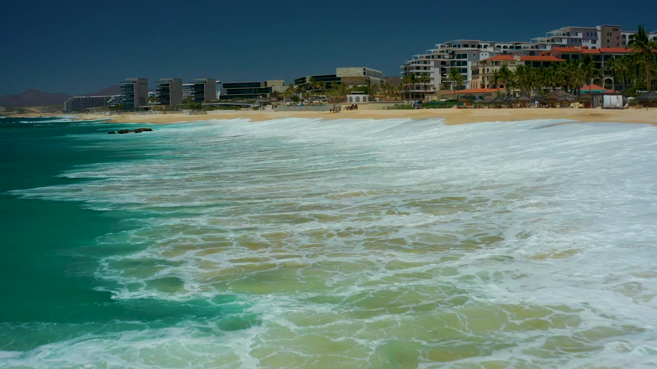 Wide shoreline view with turquoise surf, sandy beach, rocks, and beachfront buildings.