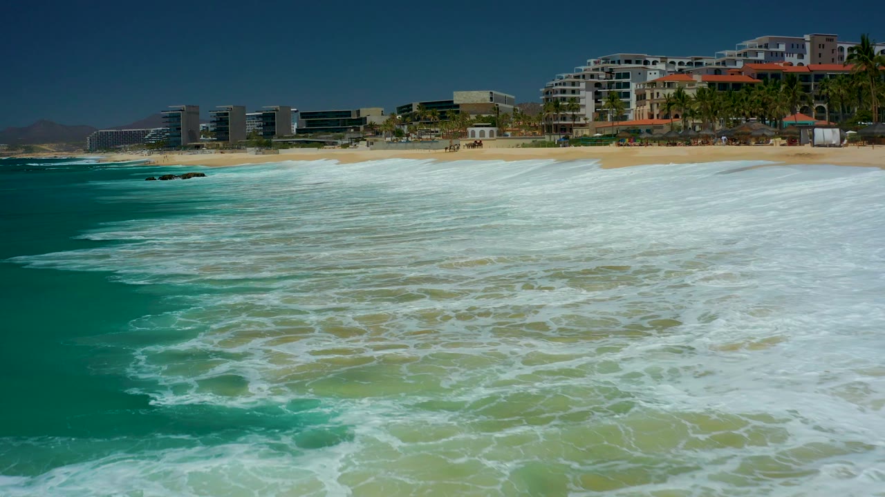 At Dreams Los Cabos in Baja California Sur, white surf advances across green-blue water toward the sand while beachfront buildings remain across the upper frame, with no visible people.