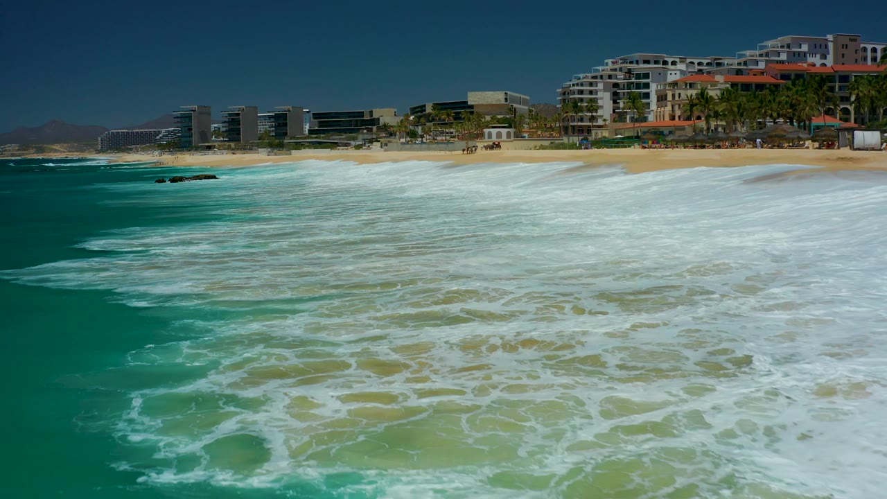 At Dreams Los Cabos in Baja California Sur, the foreground is filled by white foam over turquoise water, with sand and modern beachfront buildings across the top of the frame, with no visible people.