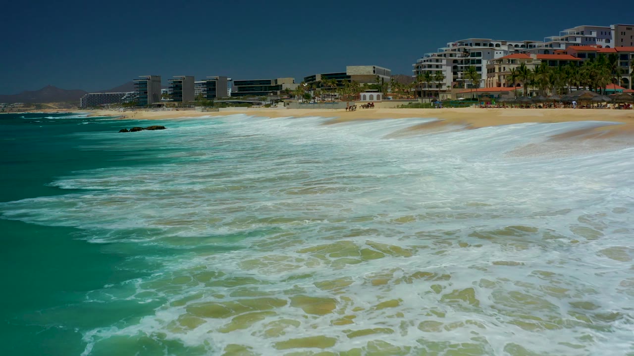At Dreams Los Cabos in Baja California Sur, breaking waves push diagonally across the turquoise water toward a sandy shore with rocks near the left center and buildings beyond, with no visible people.