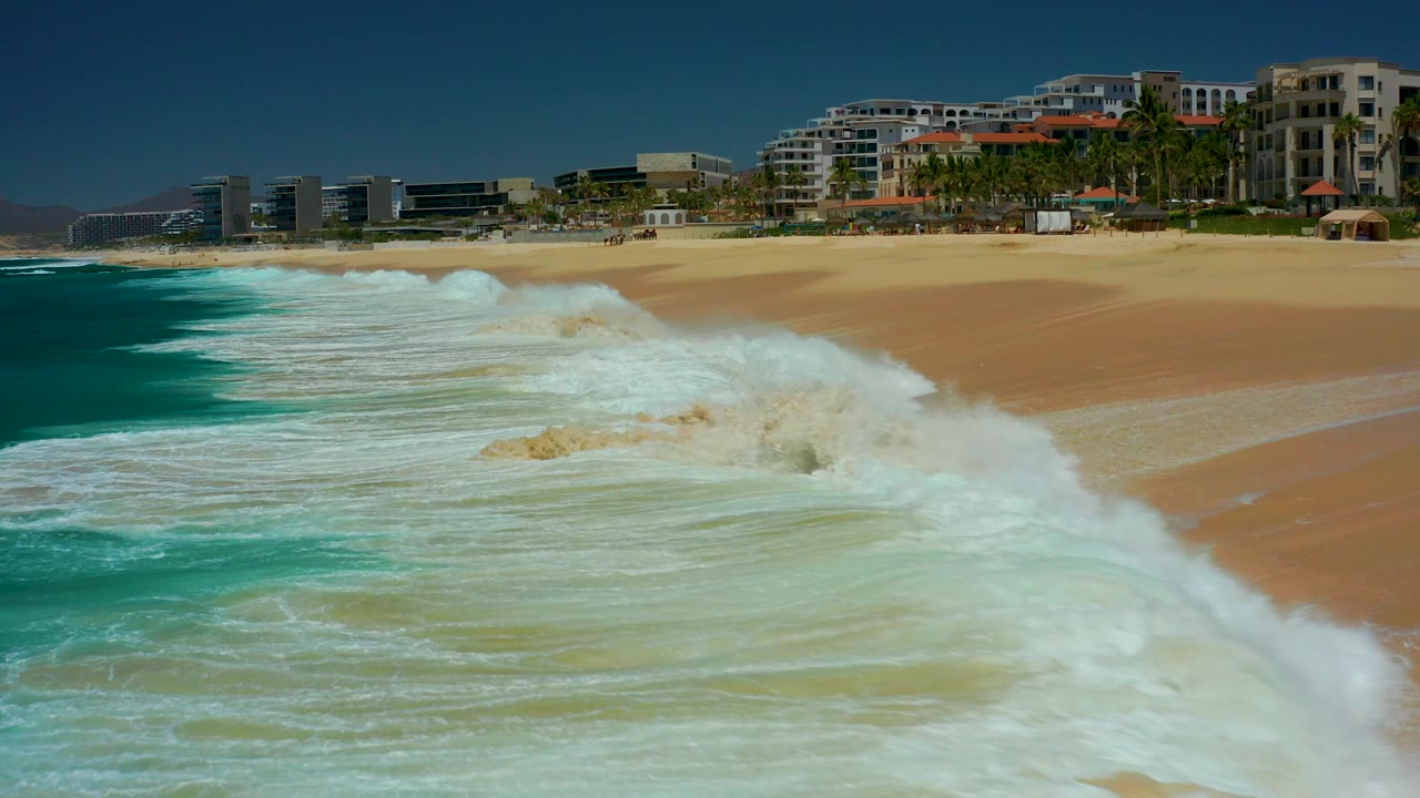 At Dreams Los Cabos in Baja California Sur, a large white wave breaks across the foreground toward a sandy beach backed by palm trees and beachfront buildings, with no visible people.
