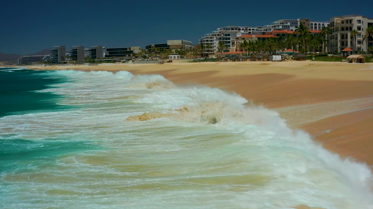 White surf spreads farther across the beach below palm trees and buildings.