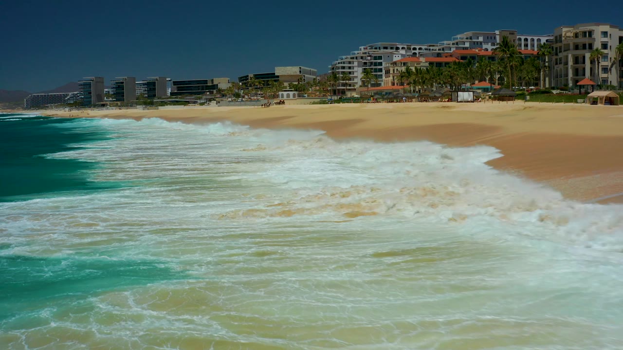 At Dreams Los Cabos in Baja California Sur, whitewater fills much of the foreground shallows beside a pale sandy beach, with palms and beachfront buildings across the top of the frame and no visible people.