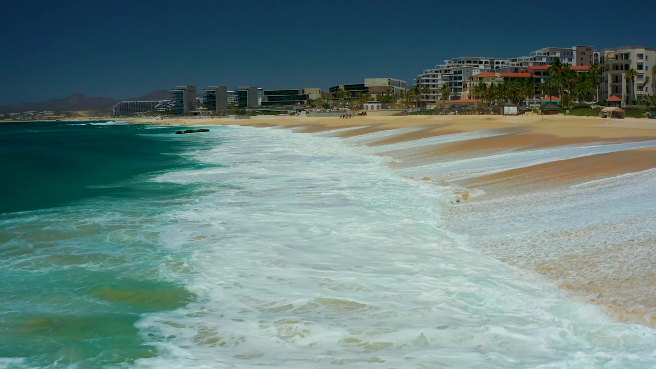 White water blankets more of the Dreams Los Cabos beach, with turquoise ocean at left, tan sand at right, beachfront buildings and palms in the distance, and no visible people.