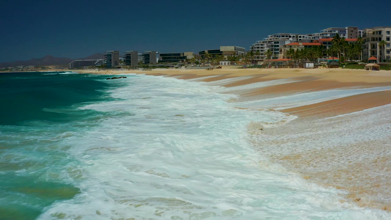 The surf pattern shifts along the Dreams Los Cabos shoreline, leaving wet streaks on the sand while blue-green water, resort buildings, palms, and distant hills remain visible, with no visible people.