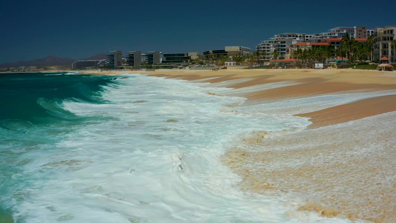 A broader sweep of the Dreams Los Cabos beach shows white water sliding up the sand, darker ocean at left, pale foam trails, resort buildings, palm trees, and no visible people.