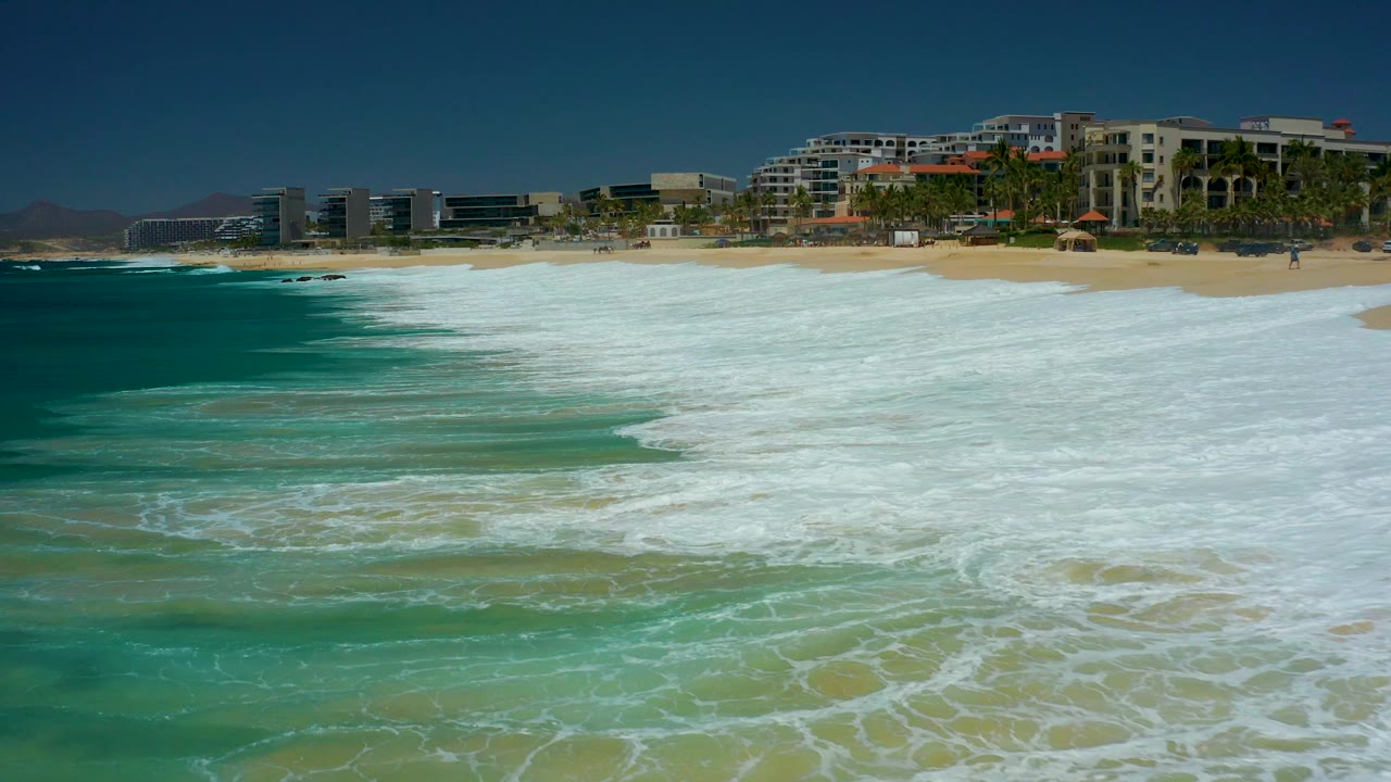At Dreams Los Cabos in Baja California Sur, white surf spreads across the sandy shoreline while turquoise water and beachfront resort buildings sit behind it, with no visible people.