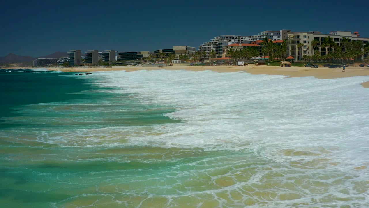 A second of surf at Dreams Los Cabos shows foamy water advancing over the sand, deeper blue ocean at left, and resort buildings with palms along the upper shore, with no visible people.