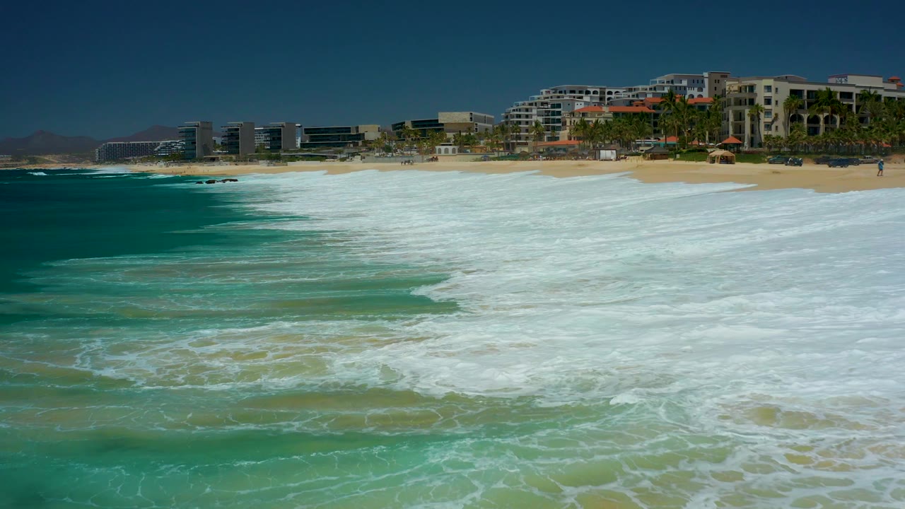 Whitewater covers more of the near shoreline at Dreams Los Cabos, with a deep blue band of ocean on the left and modern beachfront buildings across the background, with no visible people.