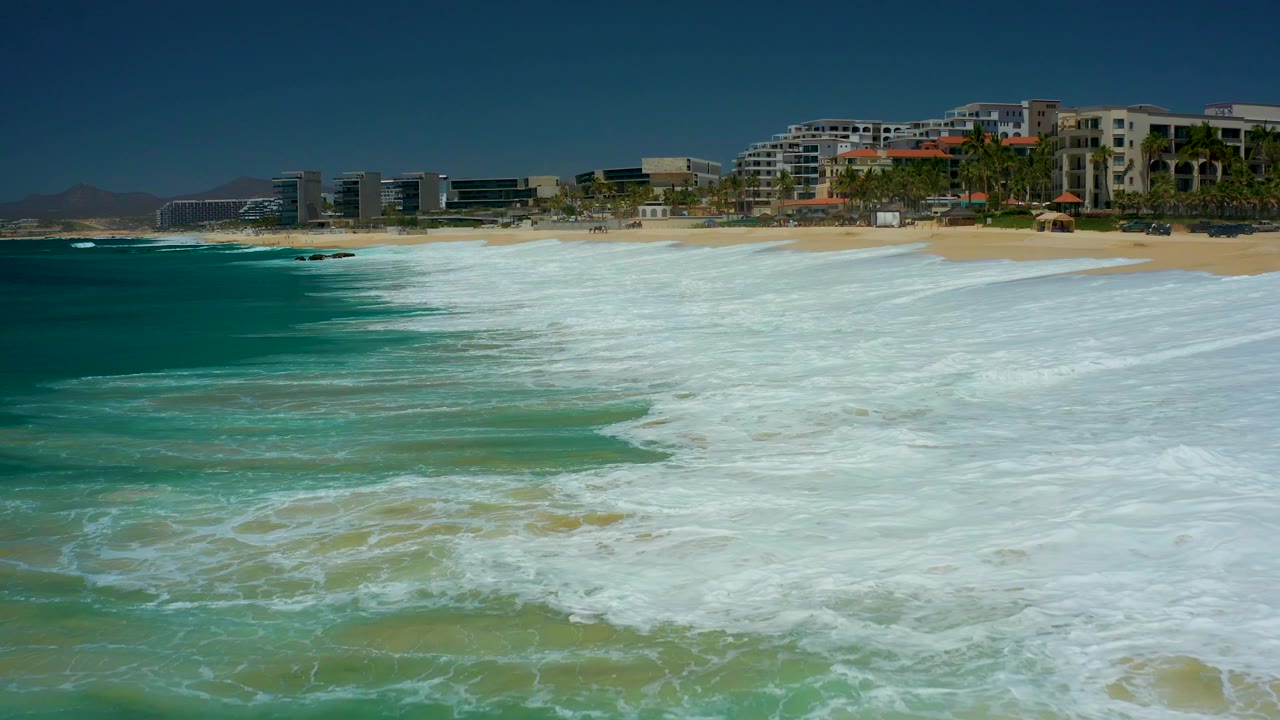 Large white wave wash crossing the foreground with beach and resort buildings behind it.