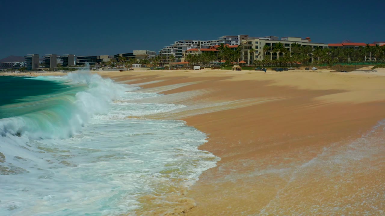At Dreams Los Cabos in Baja California Sur, white foam pushes farther up the sandy shoreline while turquoise water remains at left and the beachfront buildings stay fixed in the background with no visible people.