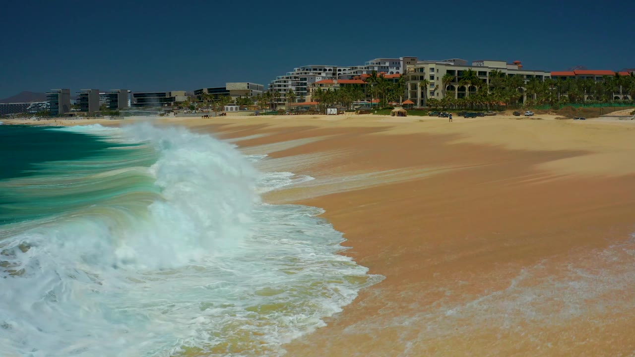 At Dreams Los Cabos in Baja California Sur, a larger white wave curls in from the left and sends foam diagonally across the beach, with resort buildings and palms beyond the sand and no visible people.