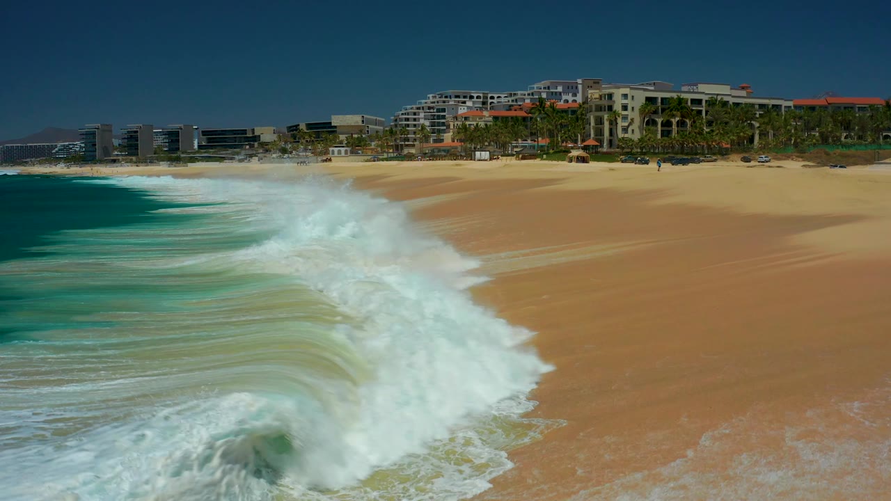 At Dreams Los Cabos in Baja California Sur, broken surf spreads in a long white band across the lower frame, with open sand, palm trees, and resort buildings behind it and no visible people.