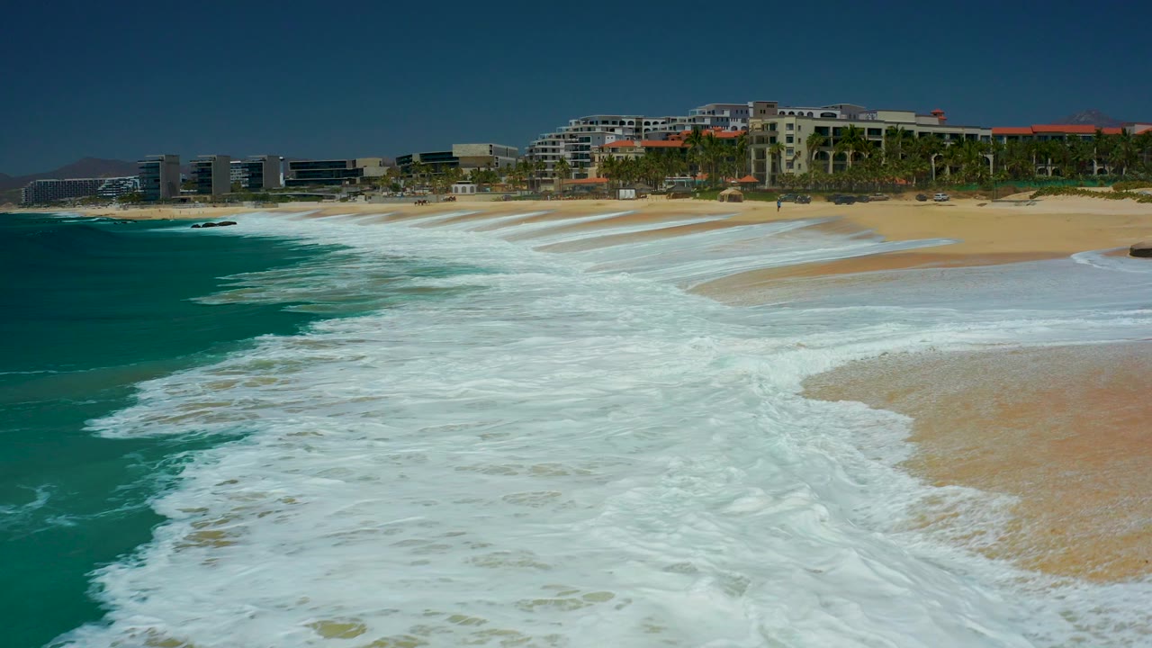 At Dreams Los Cabos in Baja California Sur, white surf fills the lower frame as turquoise water breaks onto tan sand, with resort buildings and palm trees in the background and no visible people.