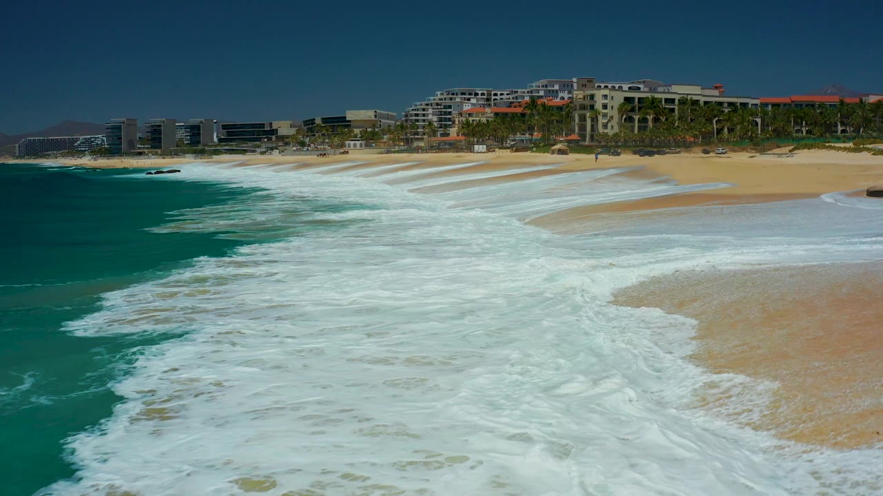 At 0:01, the foamy wash reaches farther across the foreground sand while turquoise water remains on the left and resort buildings line the background at Dreams Los Cabos, with no visible people.