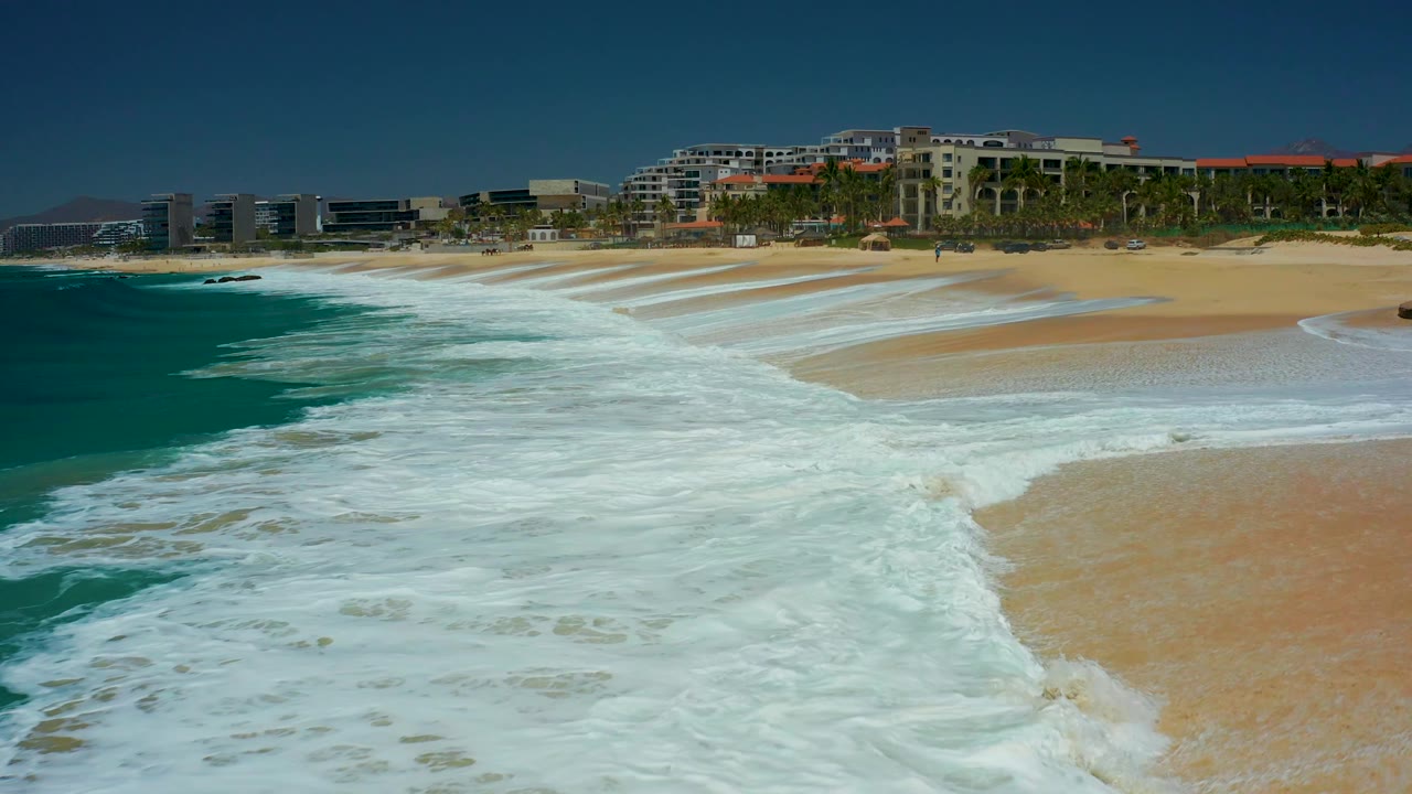 At Dreams Los Cabos, a curved line of white foam cuts across the beach as the ocean stays turquoise on the left and resort buildings with palms sit beyond the sand, with no visible people.